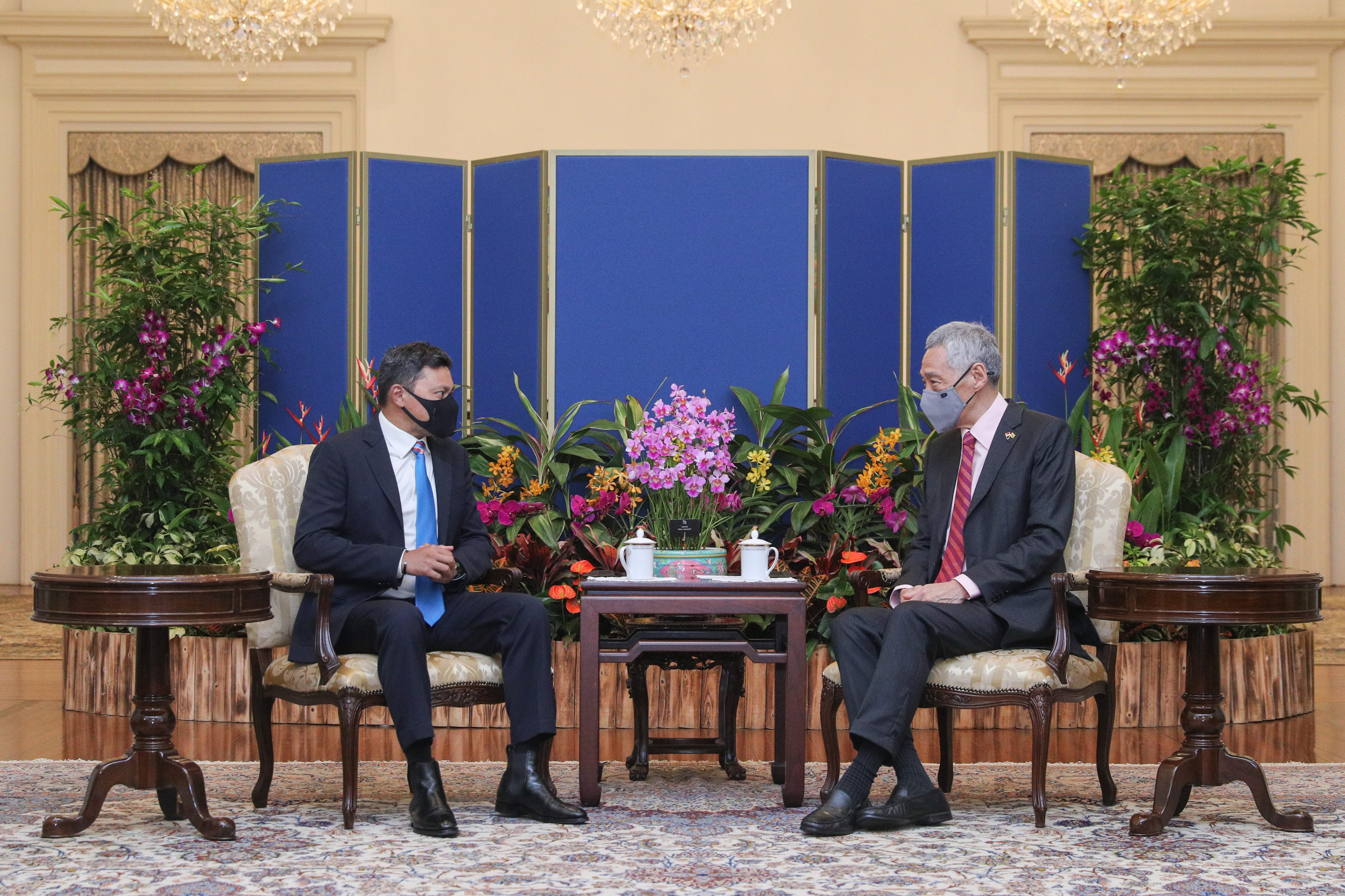 Two men in suits and face masks seated in ornate chairs across a table with floral arrangements.
